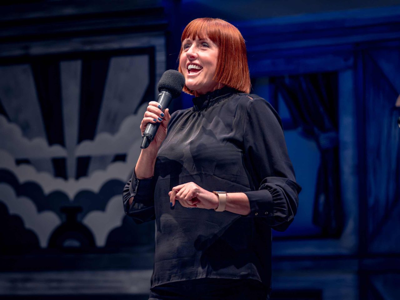 A woman with an orange bob is holding a micropho, smiling whilst addressing an audience.
