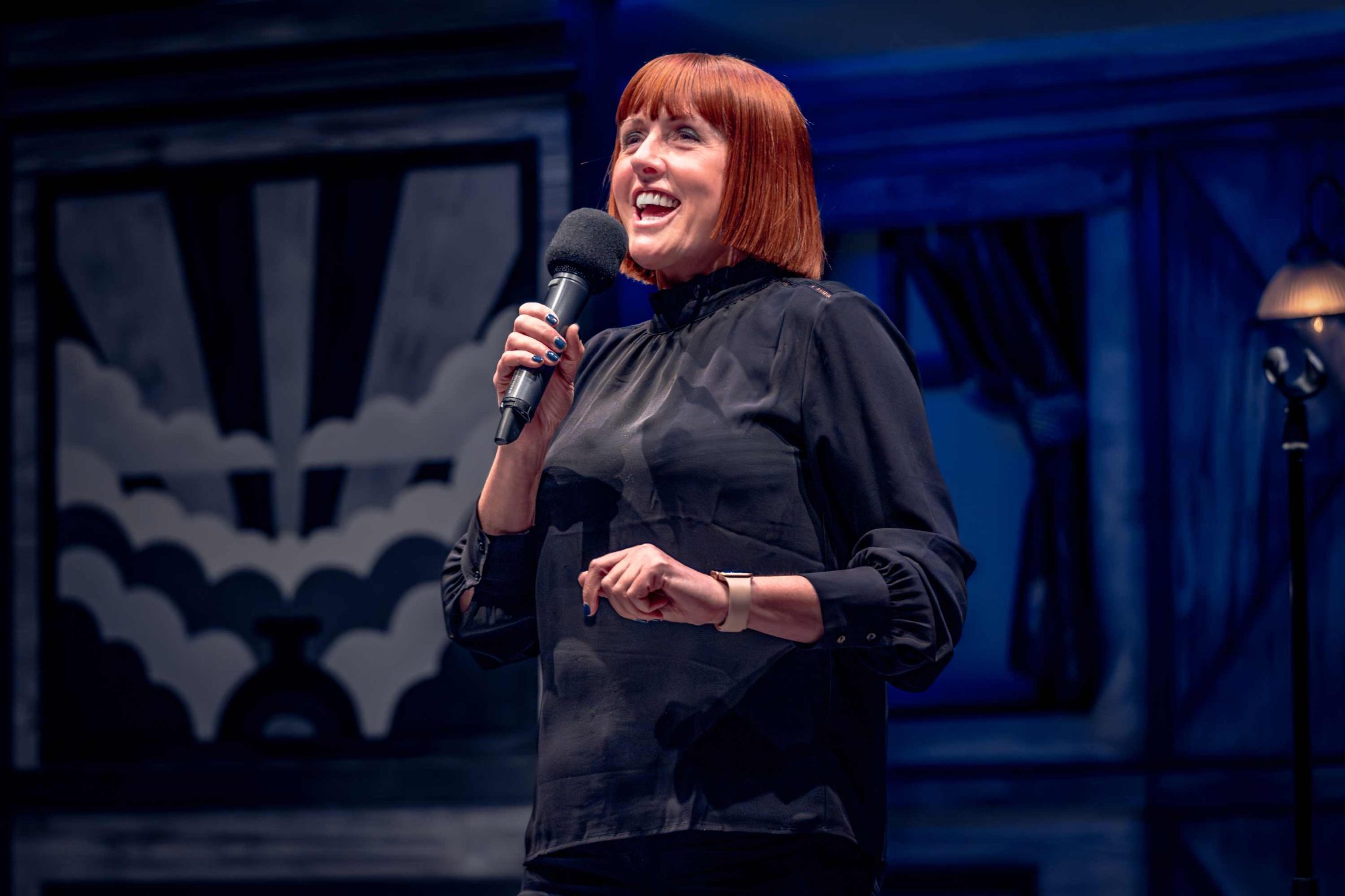A woman with an orange bob is holding a micropho, smiling whilst addressing an audience.