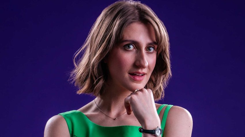 A woman with short hair and a green top smiles at us with her hand resting at her chin.