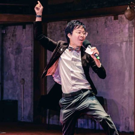 A man in a blazer jumps up, holding a microphone while his other hand waves above his head.