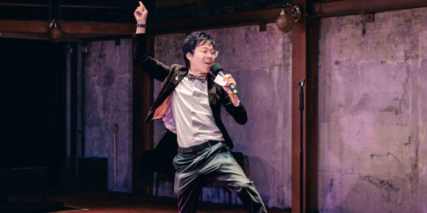 A man in a blazer jumps up, holding a microphone while his other hand waves above his head.