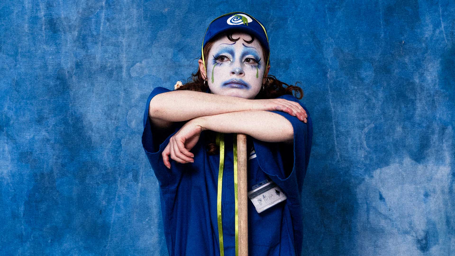 A person with clown make-up leans on a broom. They look sad, and they are wearing the uniform of a cinema employee.