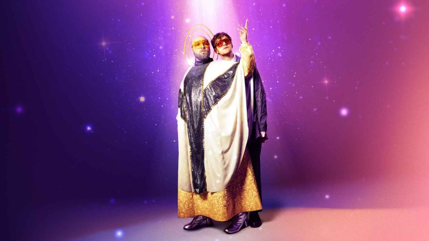 Two people wearing priest's robes stand light coming down from above. The background is purple and sparkling.