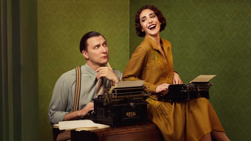 A man and a woman sit at a table dressed in 1920s clothes. They smile, both with typewriters on their laps.