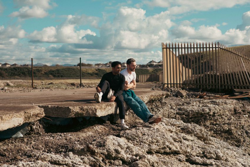 Two young men sit on a slab of concrete in an are of wasteland. Behind them are metal fences and open green spaces.