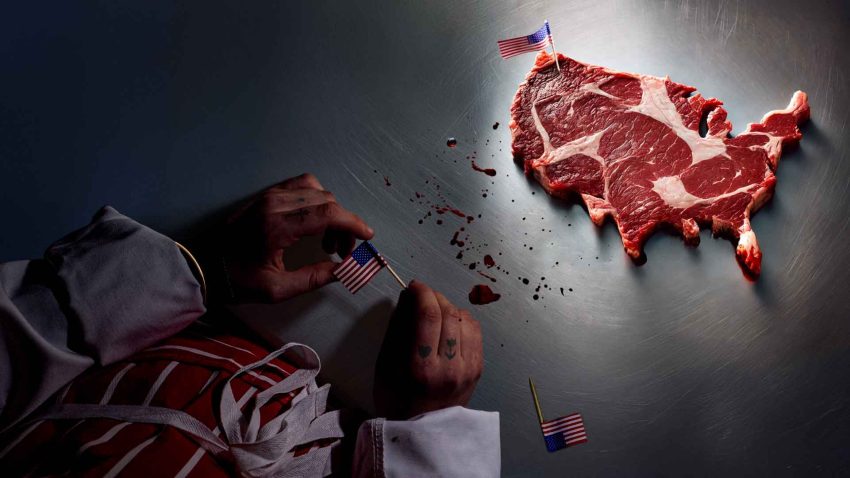 The hands of a person in a chefs uniform can be seen holding a small American flag. On the table is a steak in the shape of the USA with a small American flag stuck in it.