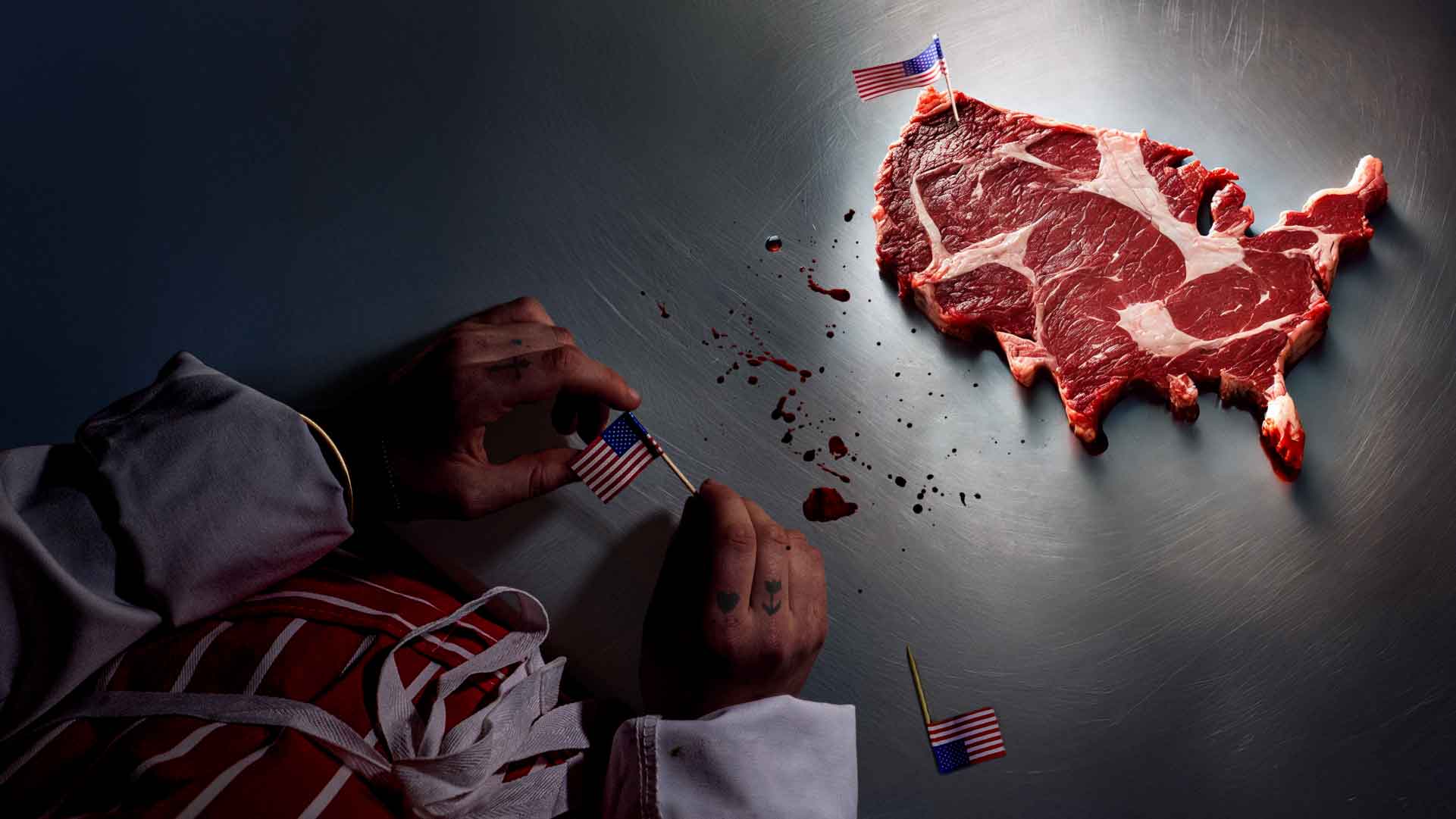 The hands of a person in a chefs uniform can be seen holding a small American flag. On the table is a steak in the shape of the USA with a small American flag stuck in it.
