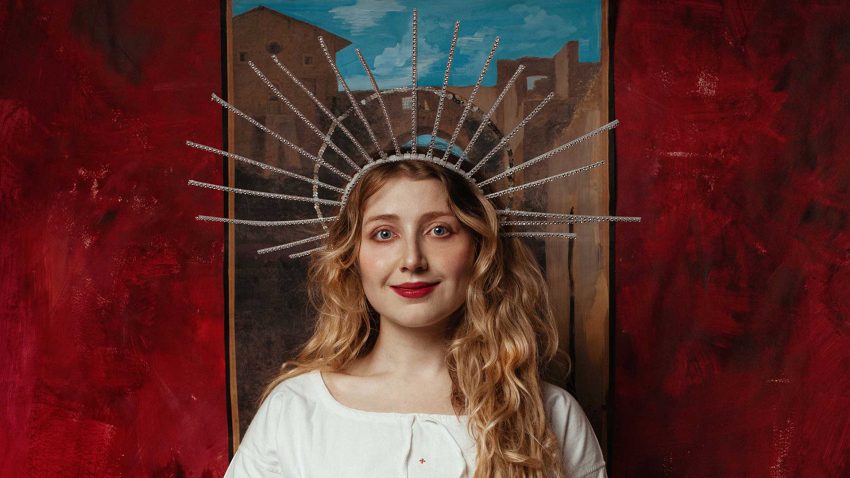 Bebe Cave wears a white shirt and a large halo-like headpiece. She smiles.