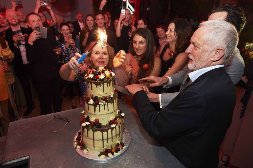 Jeremy Corbyn cutting a three layer cake while a sparkler is lit at the top of the cake.