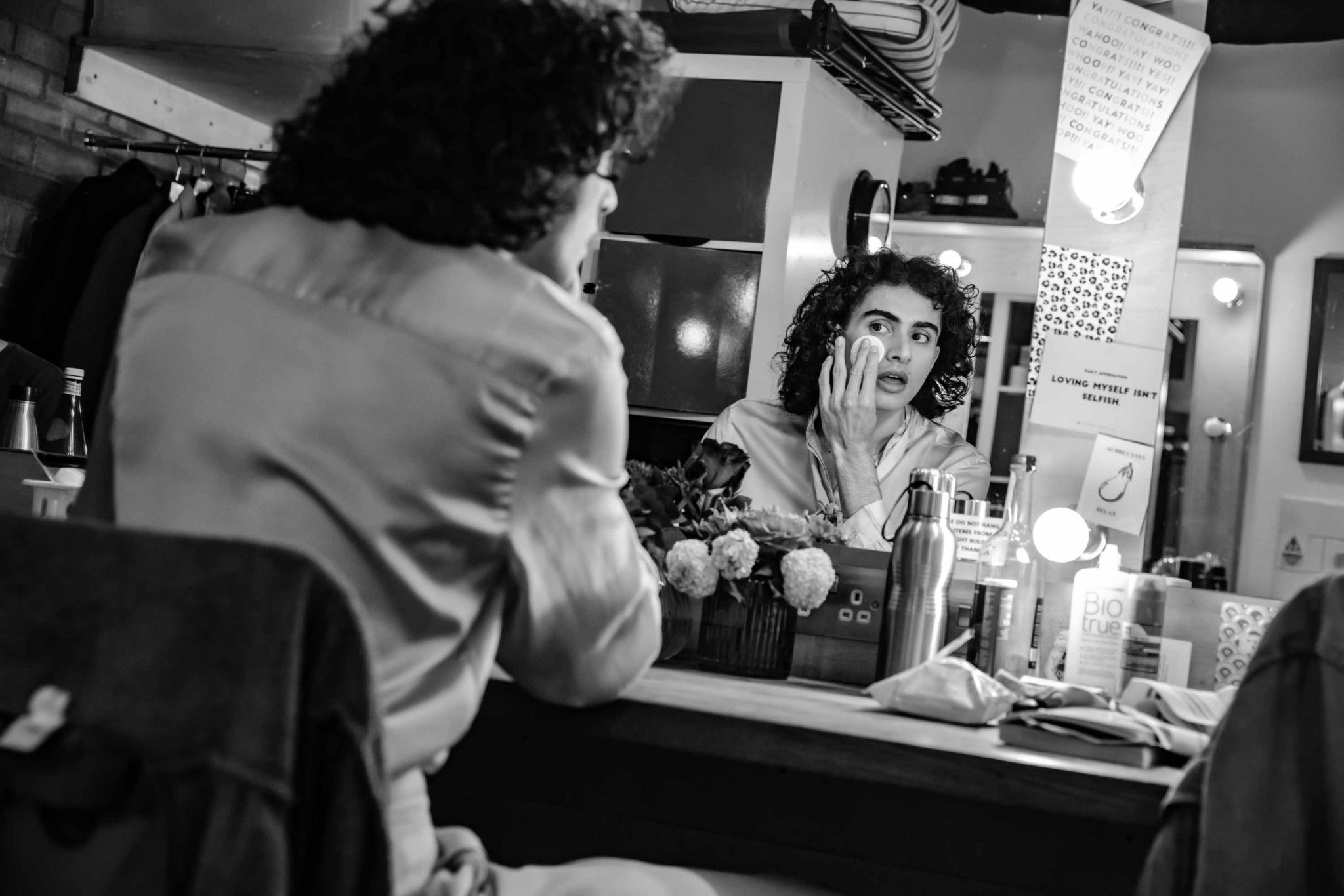 A person with curly hair looking into a backstage mirror wiping off makeup, the image is black and white