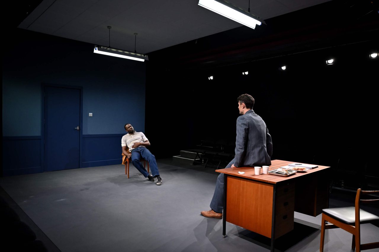 A man sits on a chair very relaxed holding his stomach in an office and another man sits facing him leaning on the office desk