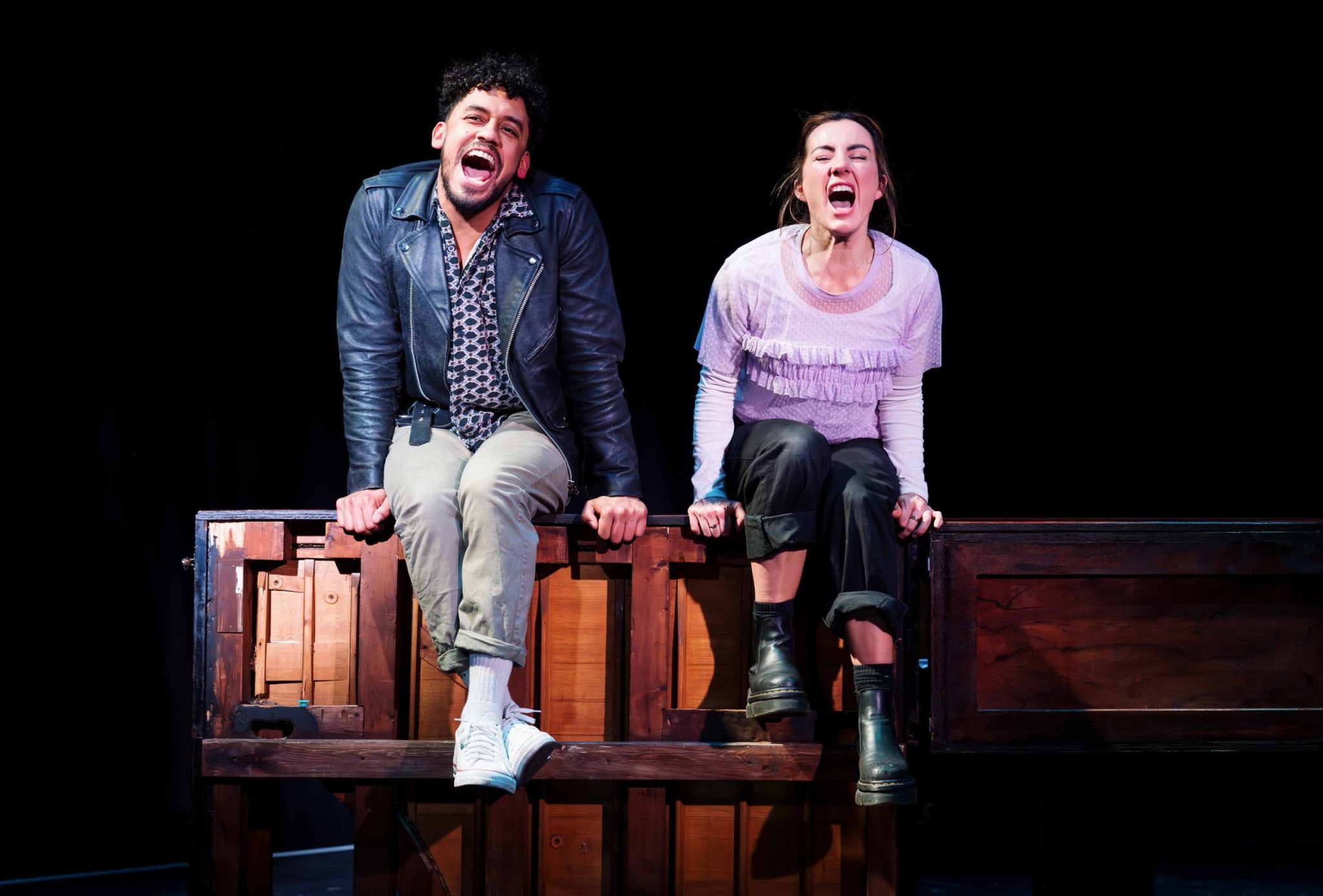 A woman in a purple shirt and black trousers and a man in a leather jacket and cream trousers sit and shout into the audience