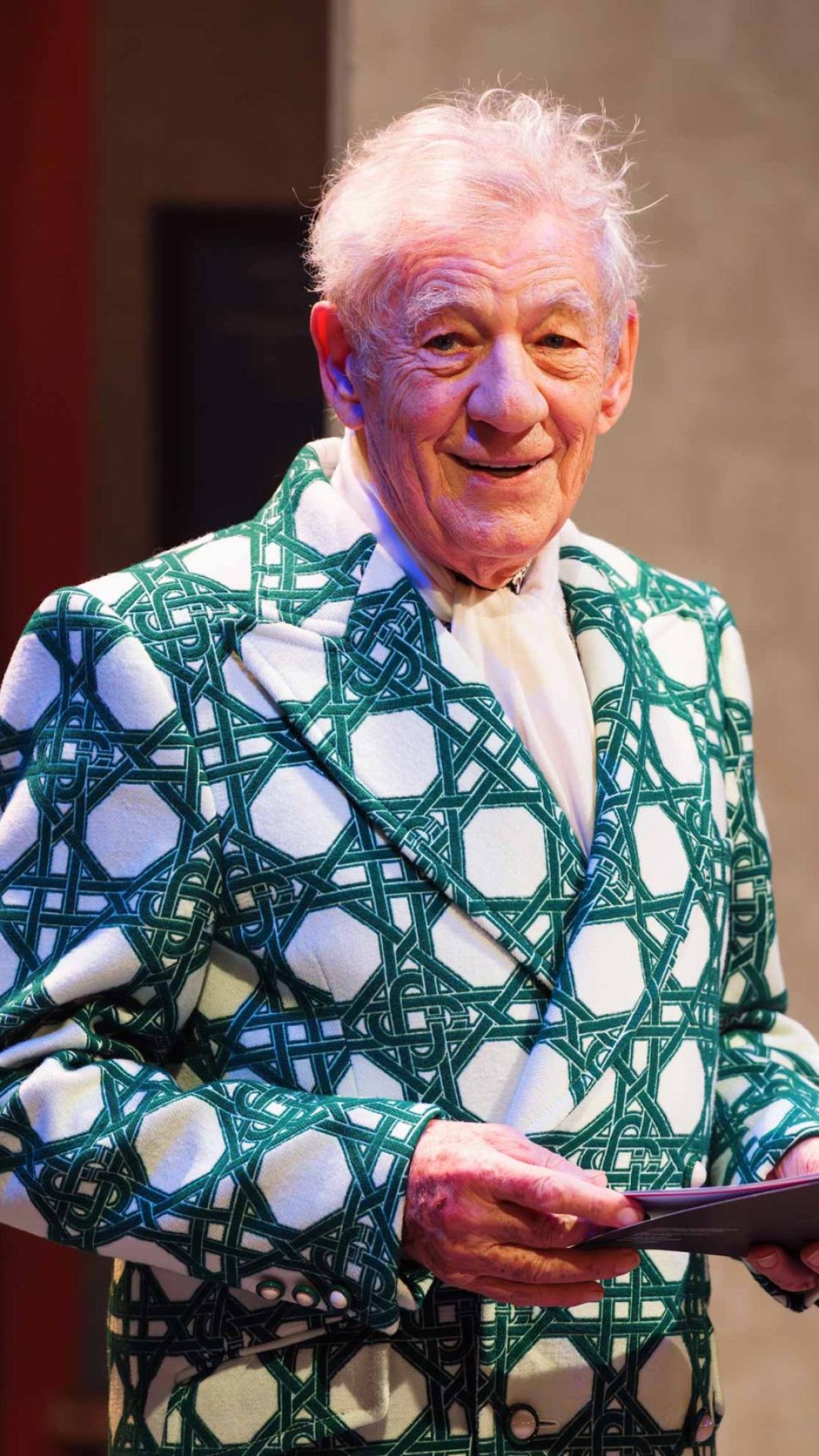 Sir Ian Mckellen in a green and white suit jacket holding a programme smiling