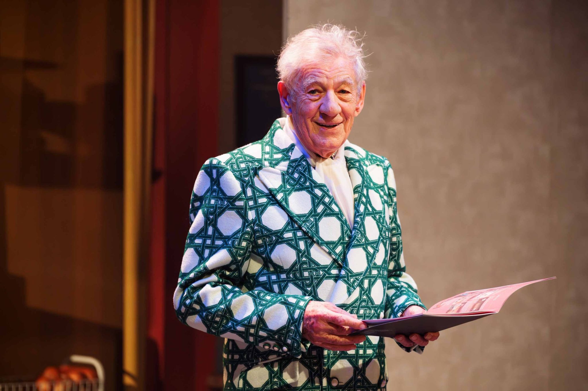 Sir Ian Mckellen in a green and white suit jacket holding a programme smiling