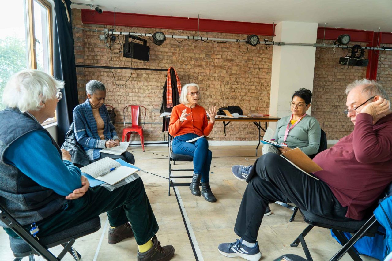 An image of a Creative Engagement group sitting and discussing poetry, five people sit in a circle on chairs with pens and notepads