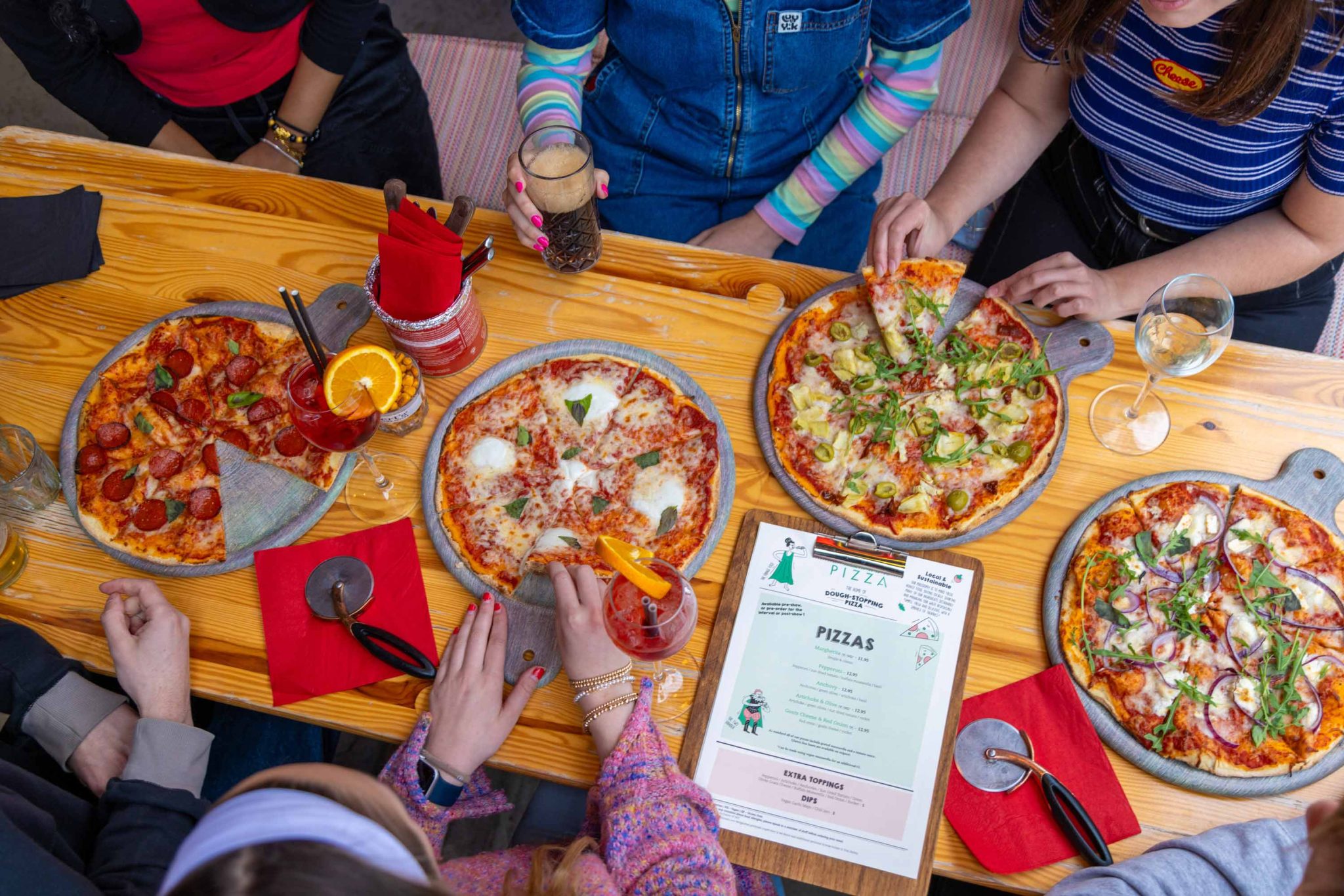 An image of a table with 4 Pizza's, drinks and 6 people sitting around the table