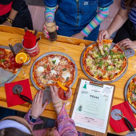 An image of a table with 4 Pizza's, drinks and 6 people sitting around the table