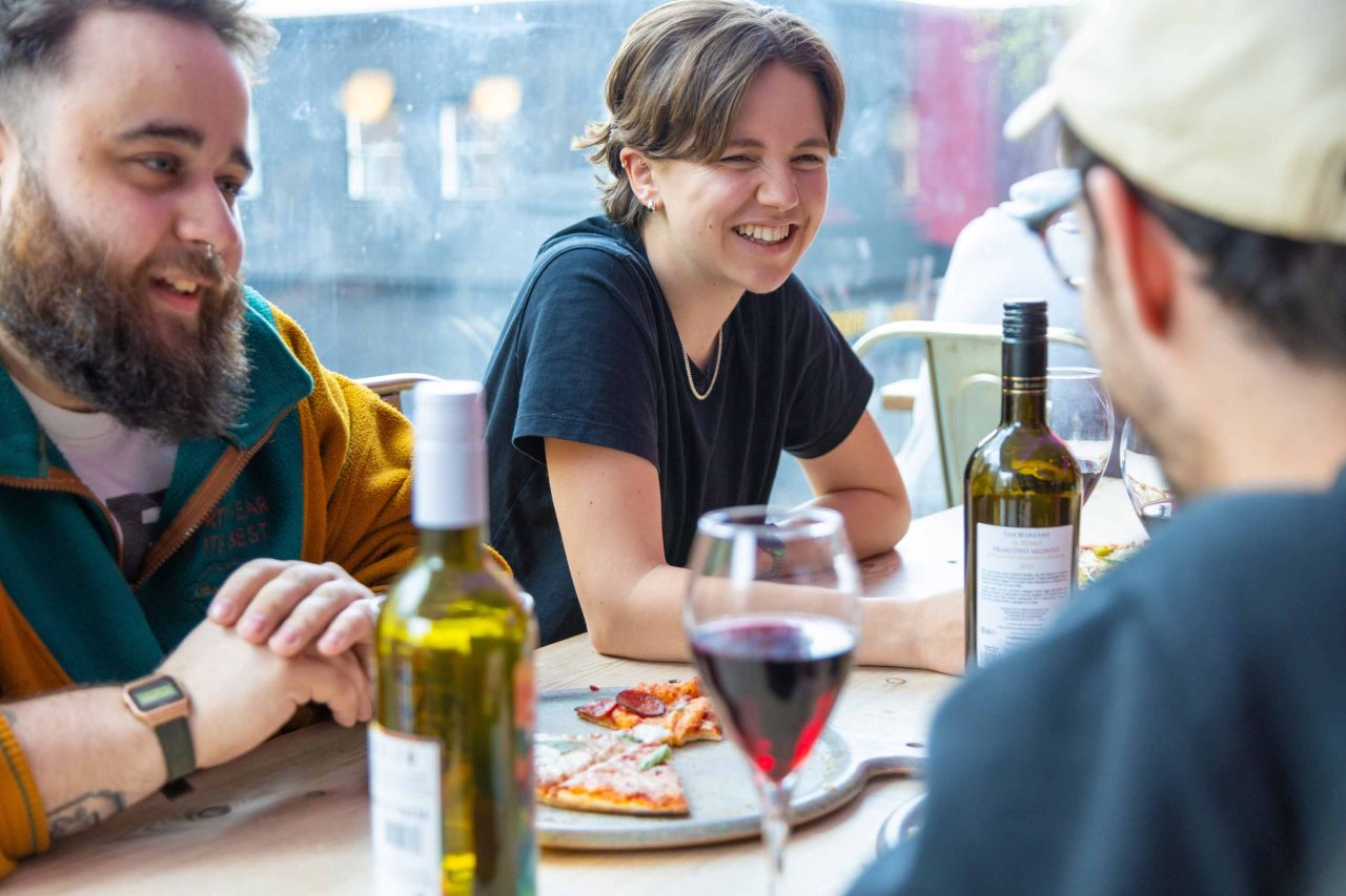 Photo of people enjoying pizza and wine, 3 friends smiling at each other a table in casual clothing