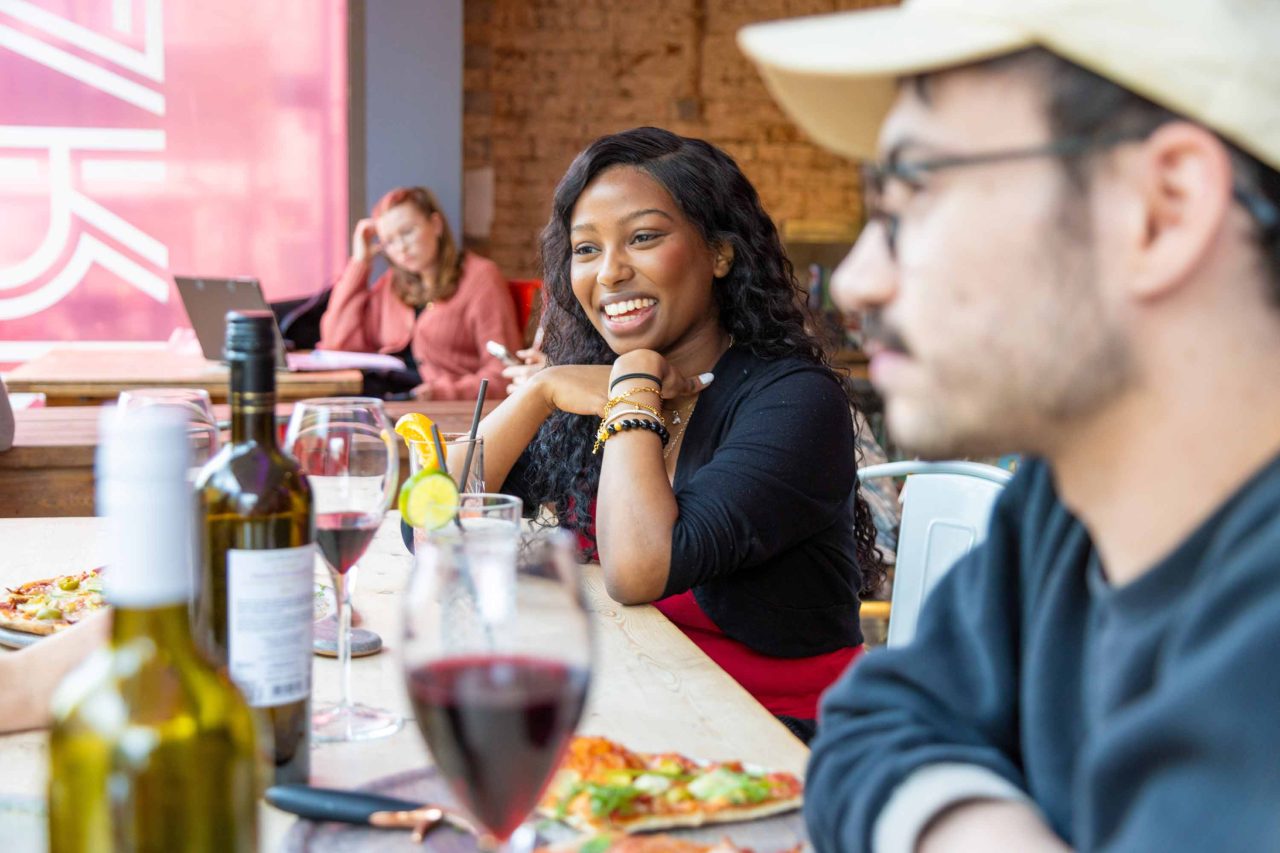 Photo of people enjoying pizza and wine, a woman in a red shirt smiles across a table, sitting beside a man in a hat