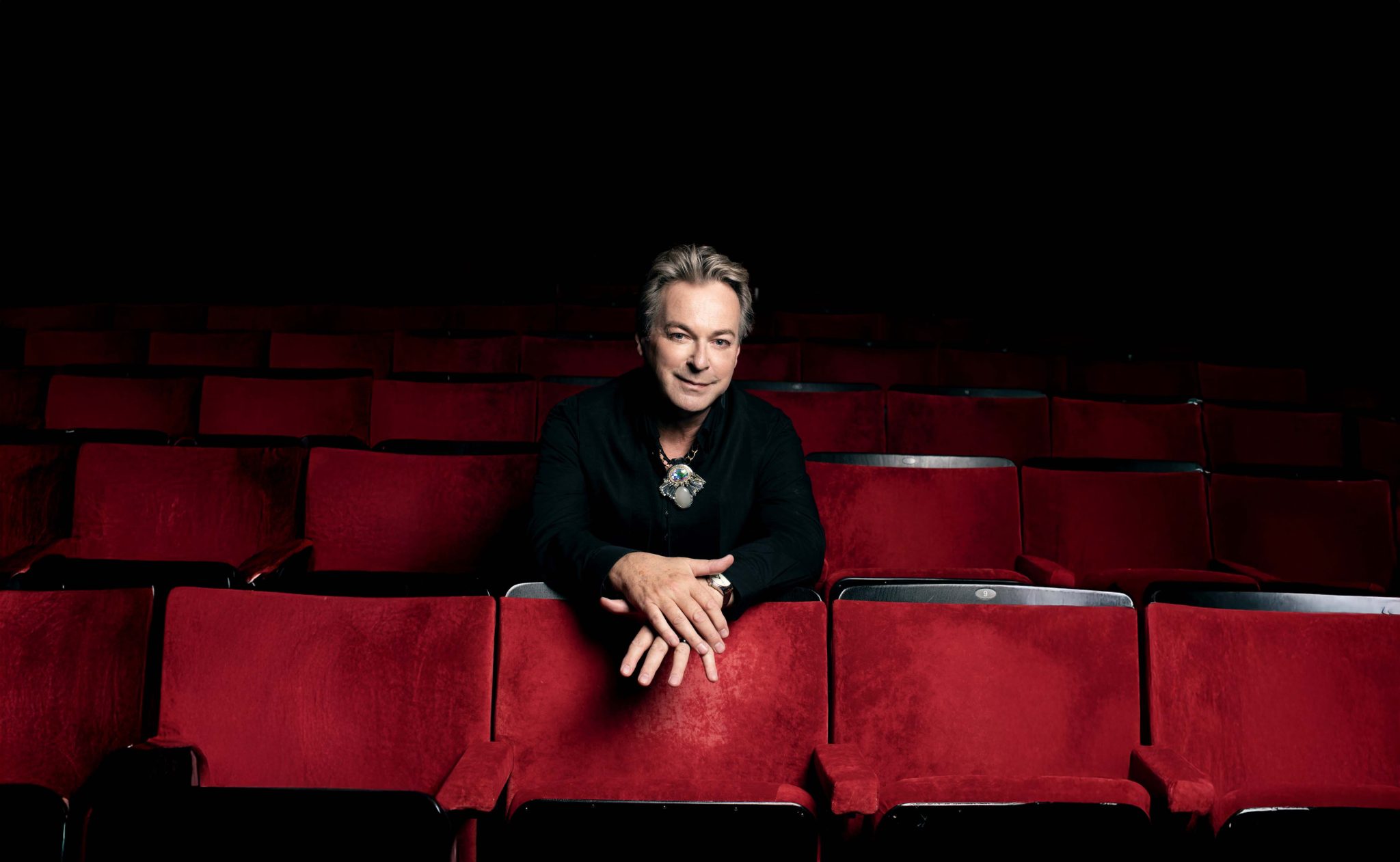 Julian Clary sits in a theatre auditorium. The chairs are plush red. Julian leans on the chair in front of him, smiling.