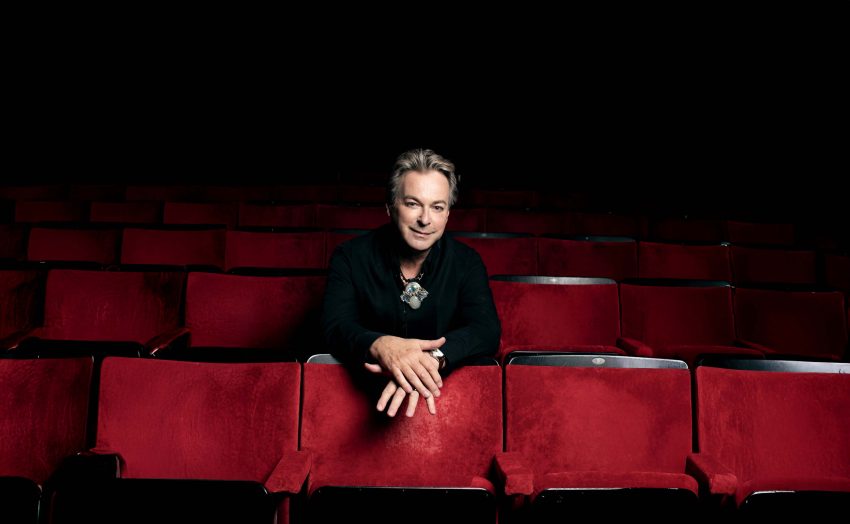 Julian Clary sits in a theatre auditorium. The chairs are plush red. Julian leans on the chair in front of him, smiling.