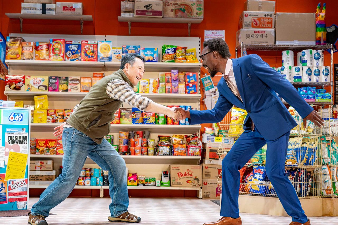 Two men shake hands enthusiastically while standing in a convenience store.