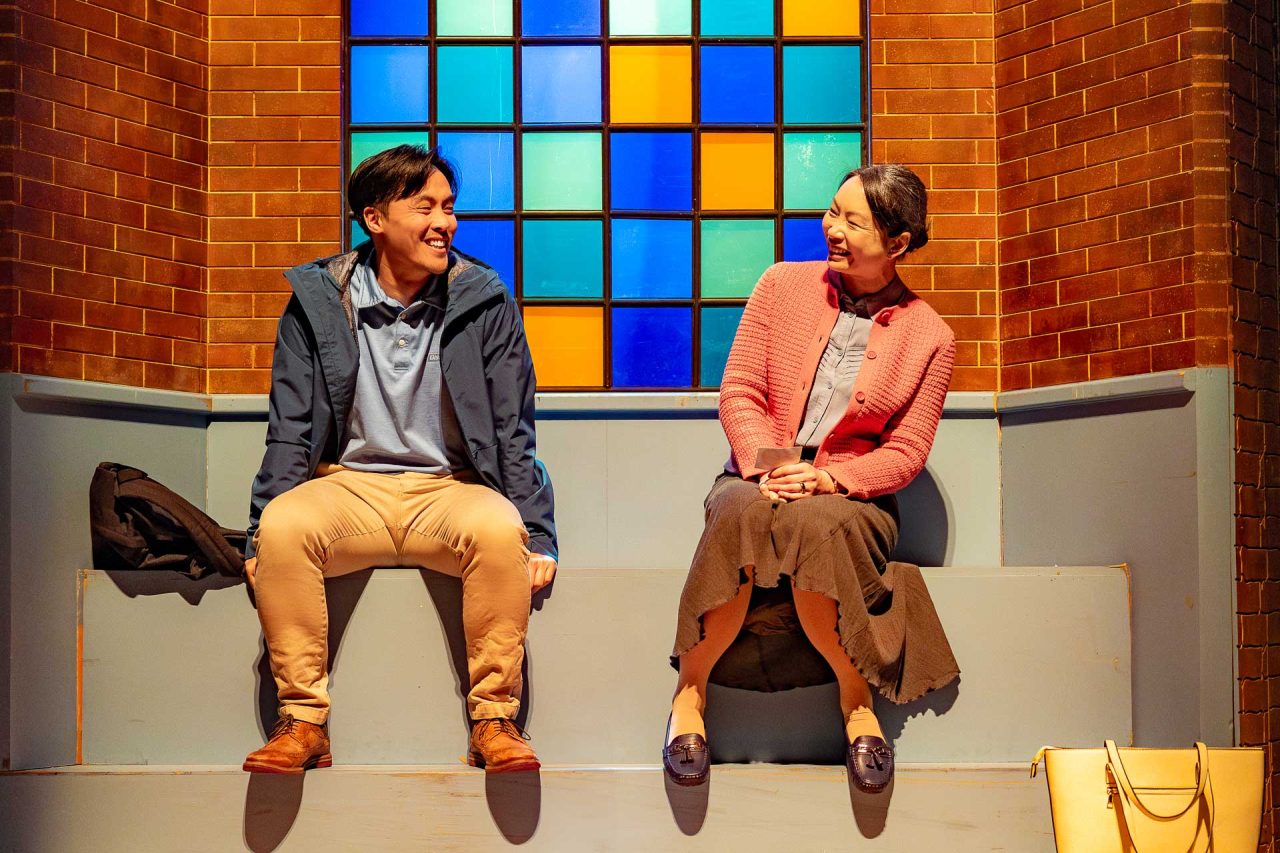 A man and a woman sit on a bench with a stained glass window behind them. They laugh with one another.