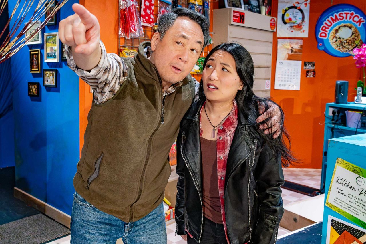 A man and a woman stand in a convenience store. The man points at something in the distance with his arm round the woman's shoulder. She looks at him with confusion.